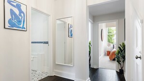 Un couloir doté d’un plancher en bois foncé, de murs blancs et d’une porte donnant sur une salle de bain avec une baignoire blanche et un espace de douche carrelé.