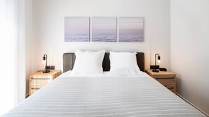 Une chambre d’hôtel avec un grand lit, deux tables de chevet avec des lampes et trois tableaux accrochés au mur représentant un coucher de soleil sur l’océan.