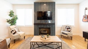 A living room with a fireplace, two striped armchairs, a coffee table, and a TV mounted above the fireplace. - Luxury East Nashville Townhome | Walk to Five Points | Dual King Suites & Arcade Room (Nashville)