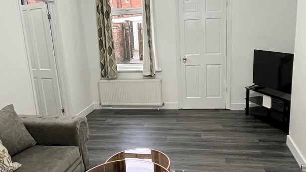Una sala de estar con un sofá gris, una mesa de centro de vidrio, un televisor sobre un mueble bajo y un radiador debajo de una ventana.