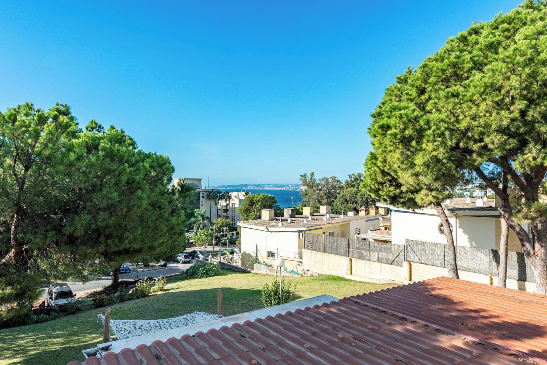 A residential area with a view of the sea, featuring houses with red-tiled roofs and green trees.