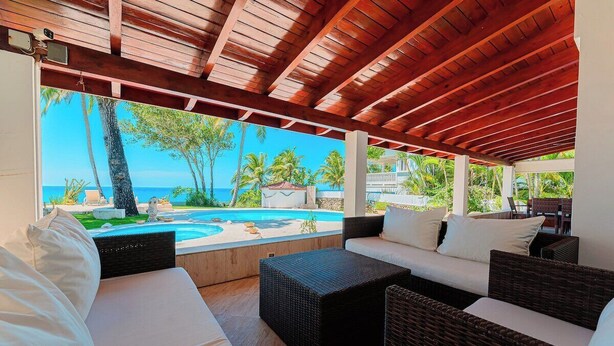 Un espace extérieur aménagé, abrité et doté de mobilier en osier, offrant une vue sur une piscine et une plage.