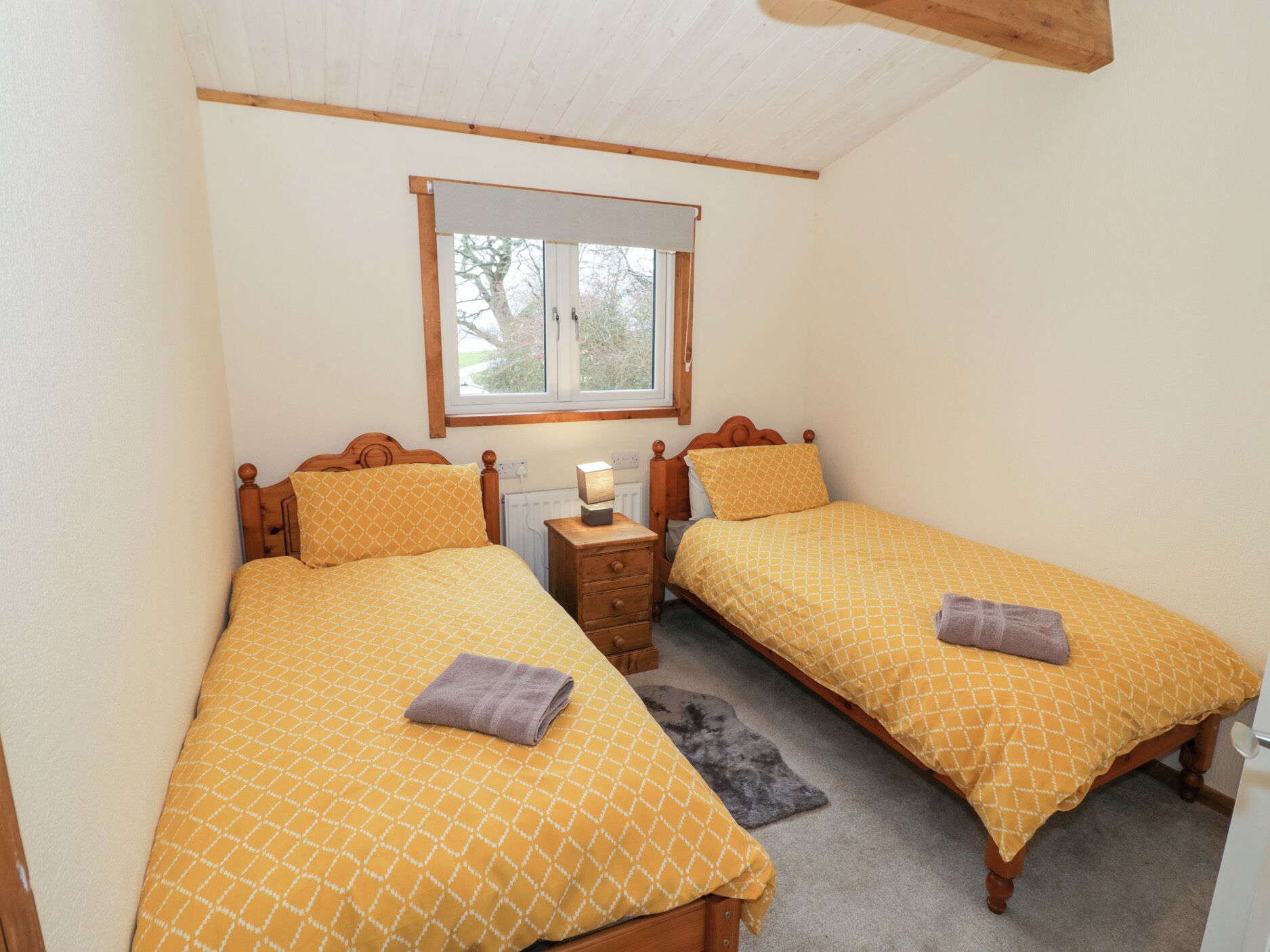 A bedroom with two single beds, each with a wooden headboard, a bedside table, and a lamp. There is a window with a view of trees and a white ceiling.