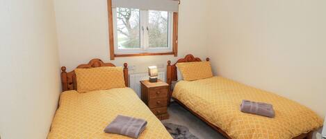 A bedroom with two single beds, each with a wooden headboard, a bedside table, and a lamp. There is a window with a view of trees and a white ceiling.