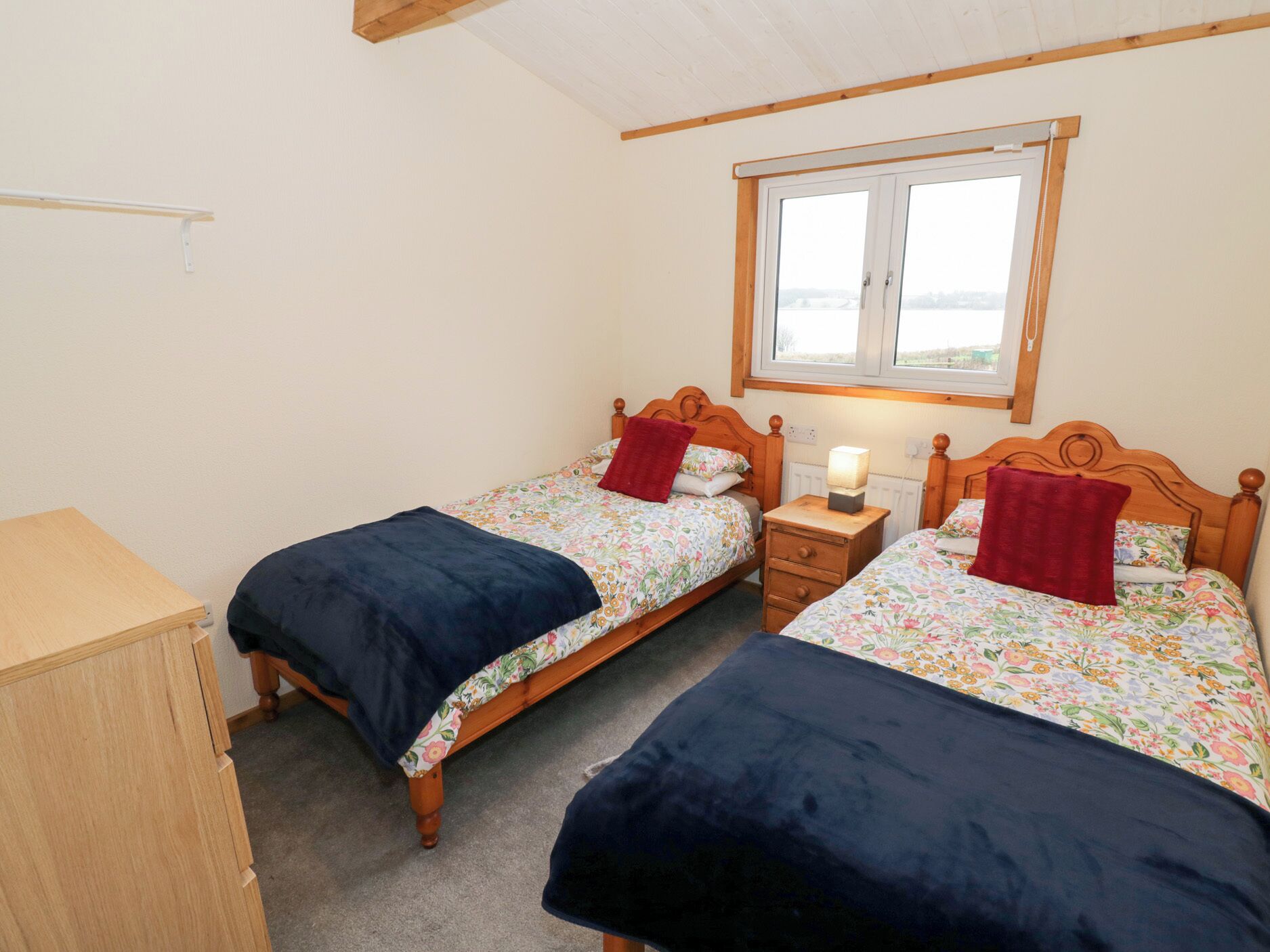 A bedroom with two single beds, wooden headboards, a nightstand with a lamp, and a window with a view.