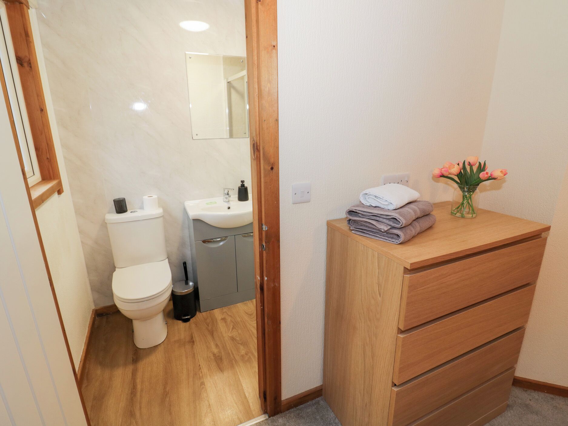 Une salle de bain comprenant des toilettes, un lavabo, un miroir et un meuble en bois contenant des serviettes et des fleurs.