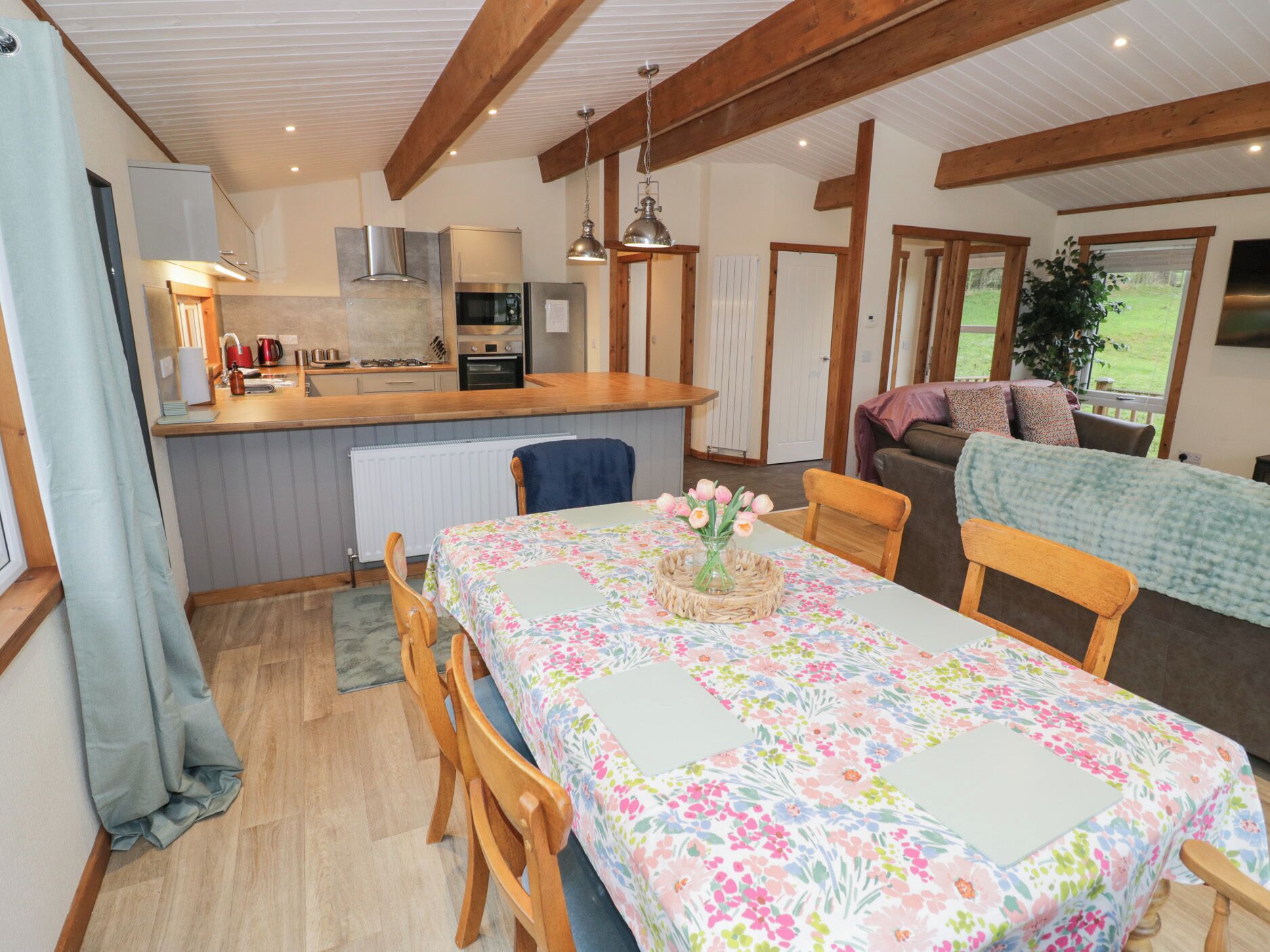 A cozy kitchen with wooden beams, a dining area with a floral tablecloth, and a living room with a sofa and a TV.