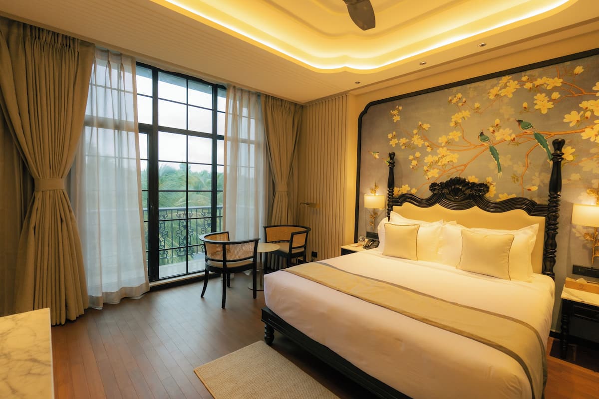 Une chambre d’hôtel avec un grand lit, une petite table avec des chaises, une vue sur la verdure par la fenêtre et un mur décoratif orné de fleurs.