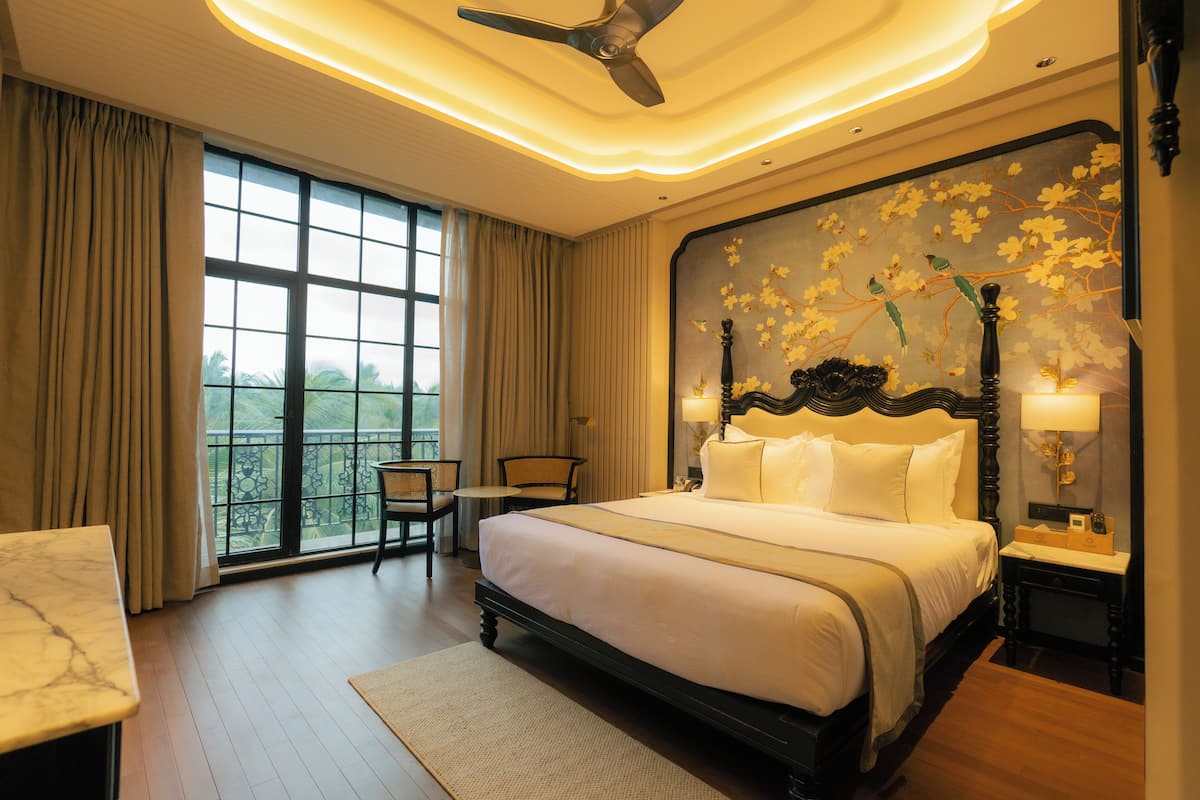 Une chambre spacieuse avec un grand lit, un ventilateur de plafond, une vue sur l’extérieur et un balcon avec une chaise.
