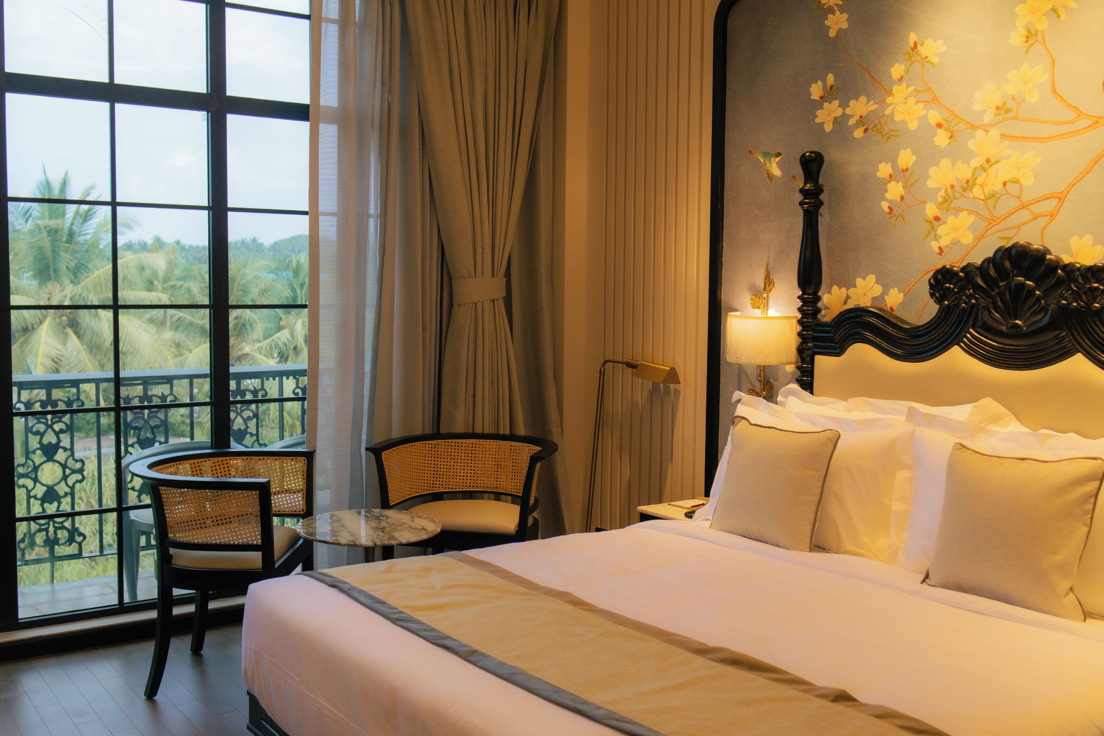 Habitación de hotel con una cama, una silla, una mesa y vistas a palmeras a través de amplias ventanas.