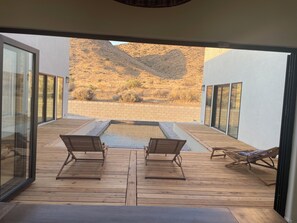 En terrasse med to træstole og en liggestol, med udsigt over en pool og et ørkenlandskab.