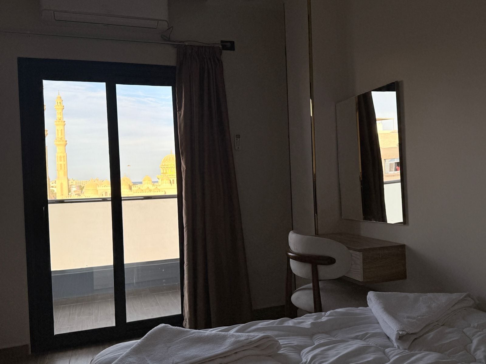 Une chambre d’hôtel comprenant un lit, un bureau, une chaise et une grande fenêtre donnant sur un bâtiment doté d’un minaret.