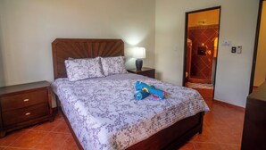 A neatly made bed with a floral bedspread and a rolled-up towel. There is a nightstand with a lamp and a small drawer. A wooden headboard is visible, and the room has a tiled floor. - Casa 78- Bright Modern Pool House with Deck (Esterillos Oeste)