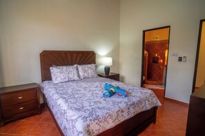 A neatly made bed with a floral bedspread and a rolled-up towel. There is a nightstand with a lamp and a small drawer. A wooden headboard is visible, and the room has a tiled floor. - Casa 78- Bright Modern Pool House with Deck (Esterillos Oeste)