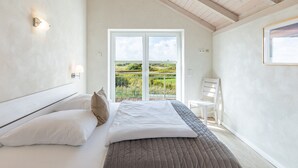 A bedroom with a bed, white walls, a view of greenery outside, and a white chair. - skandinavisches Reihenhaus in Dagbüll (Dagebüll)