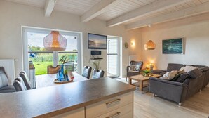 A modern living room with a kitchen area, a dining table, and a sofa. There is a television mounted on the wall and a window providing a view of the outdoors. - skandinavisches Reihenhaus in Dagbüll (Dagebüll)