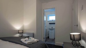 Una habitación de hotel con una cama, una mesita de noche con lámpara, un baño visible a través de una puerta abierta y una ventana pequeña.