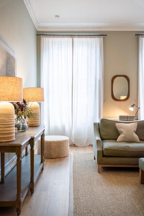 Ein Wohnzimmer mit einer Couch, einem Tisch mit Lampen, einem Spiegel und einem Fenster mit Vorhängen.