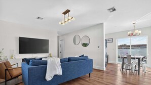 A living room with a blue sofa, a TV, a dining area with a table and chairs, and a chandelier. - Private Pool, Giant Chess, Golf, Games  by Ventur (Hollywood)