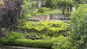 Un giardino con mura in pietra, ricco di arbusti e alberi, tra cui un salice piangente ben visibile.