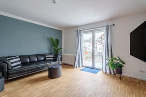 Ein modernes Wohnzimmer mit einer schwarzen Ledersofa, Holzboden, einem großen Fenster mit Vorhängen, einer Topfpflanze und einem flachbildfernseher, der an der Wand befestigt ist.
