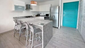 A modern kitchen with a central island, bar stools, stainless steel appliances, and a teal door. - Sanctuary Sunset -S2-209- 2 bed, 2 bath (Punta Chame)