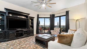 A living room with a large flat-screen TV, dark wooden entertainment unit, patterned floor tiles, a white sofa with brown cushions, and a ceiling fan. - Phoenix 6 011 (Orange Beach)