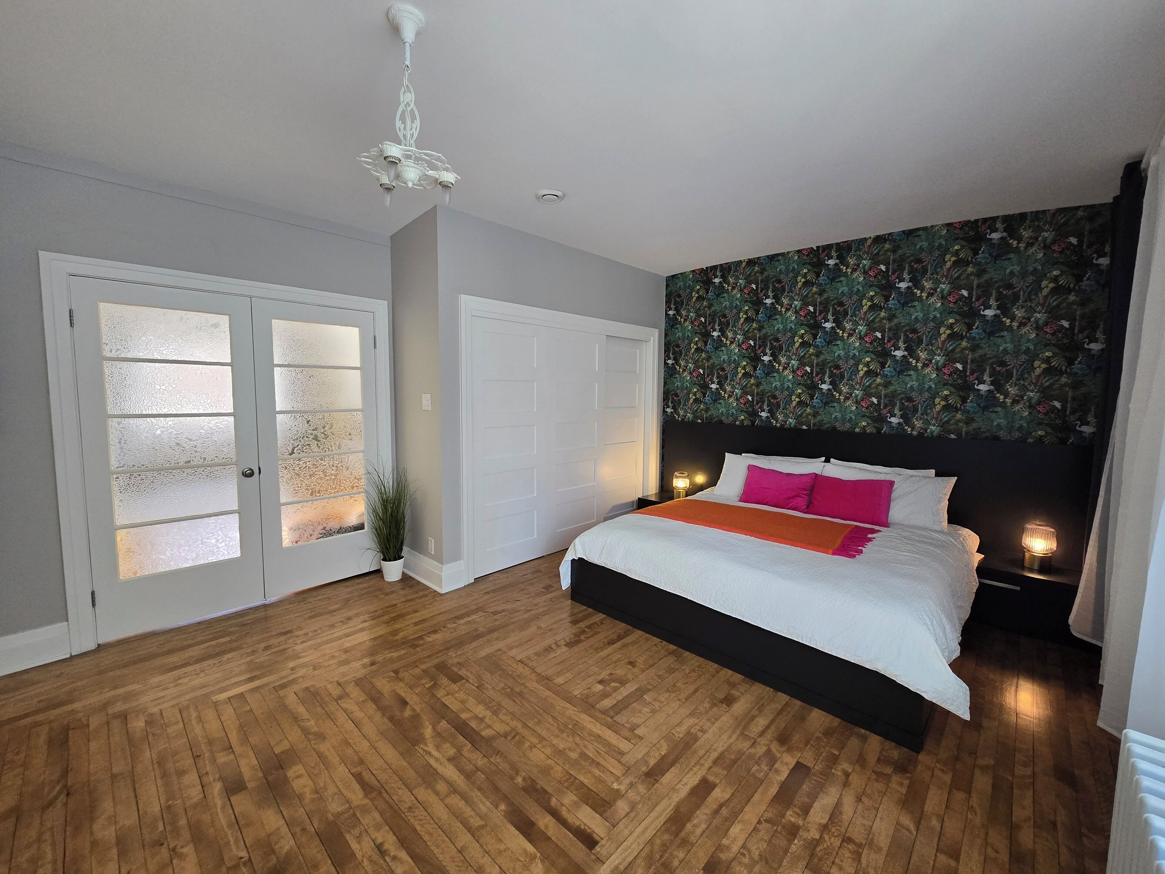 A bedroom with a wooden floor, a bed with white bedding and pink pillows, a black headboard, a floral wallpaper, and a white door with frosted glass panels.