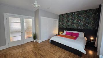 A bedroom with a wooden floor, a bed with white bedding and pink pillows, a black headboard, a floral wallpaper, and a white door with frosted glass panels.
