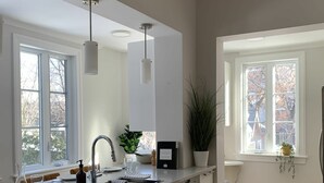 A modern kitchen with a large island, white cabinetry, a built-in dishwasher, and a window with a view of trees. - Appartements Saint-Alexandre (Longueuil)