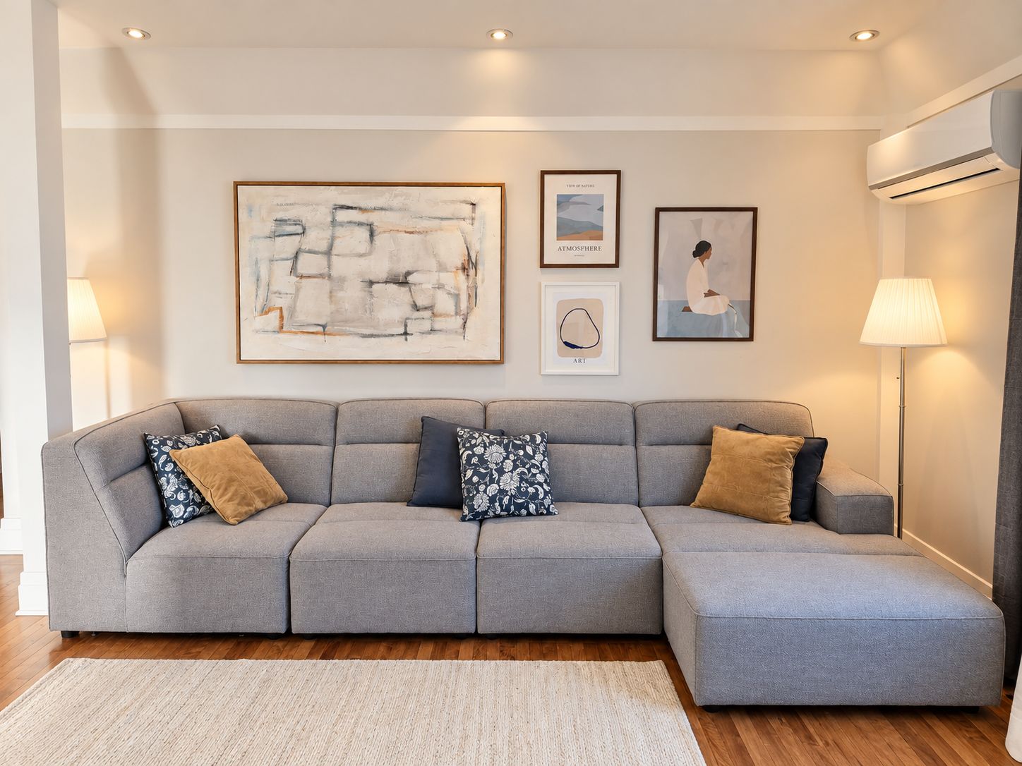 A grey sectional sofa with patterned cushions, a framed abstract painting, a framed landscape, and a lamp.