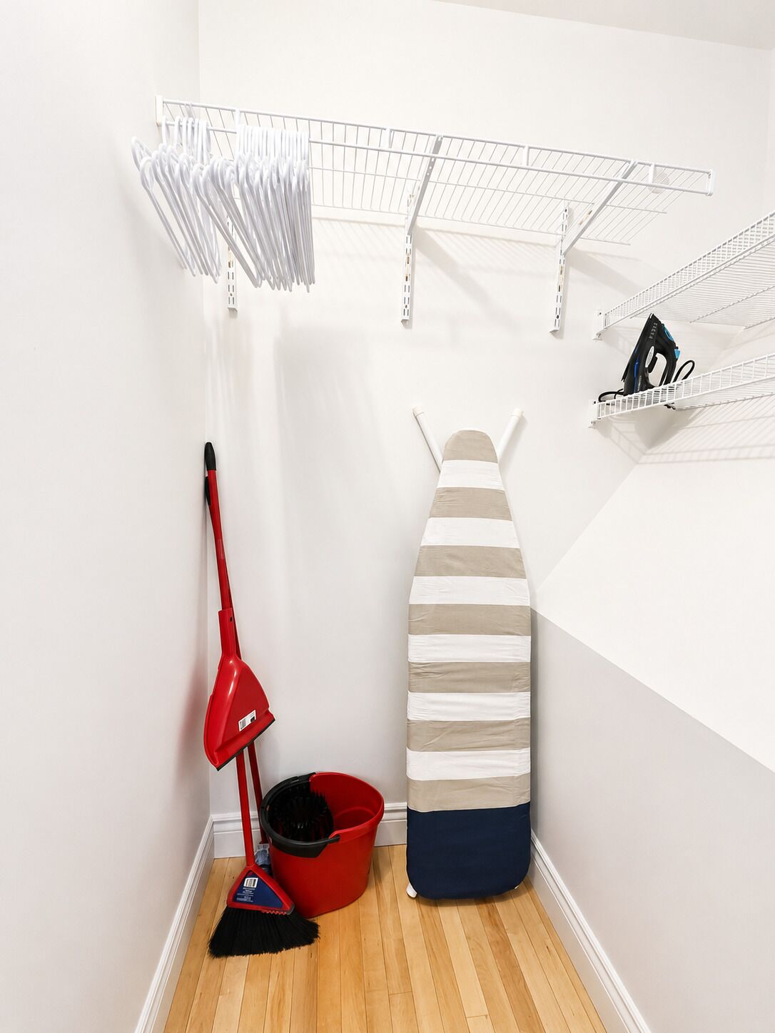 A laundry room with a red mop, a striped ironing board, a red bucket, and metal shelves for storage.