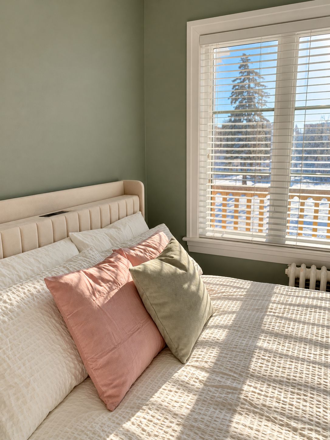 Une chambre à coucher avec un lit, des oreillers, une fenêtre avec des stores et une vue sur les arbres à l’extérieur.