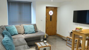 Una sala de estar con un sofá gris, cojines color turquesa, una mesa de centro de madera con una botella y vasos, una mesa de comedor de madera con fruta, y un televisor en la pared.