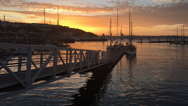Marina frente al agua al atardecer, con barcos amarrados y un puente en primer plano.