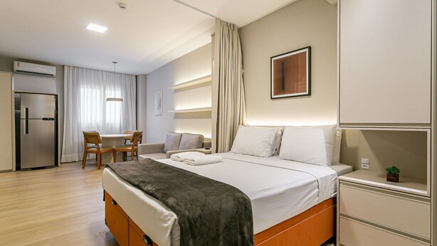Hotelkamer met een bed, nachtkastje en een kleine potplant. Er is een kitchenette met een koelkast en een eethoek met een tafel en stoelen.
