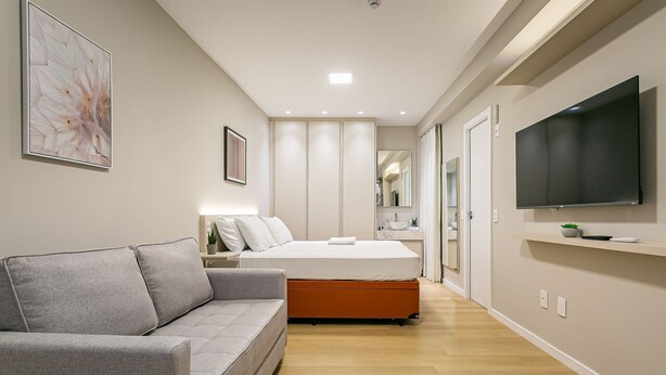 A modern bedroom with a grey sofa, a bed with an orange base, a TV mounted on the wall, and a hallway leading to a bathroom. - Walkable | Near Beiramar Mall #TM103 (Florianópolis)