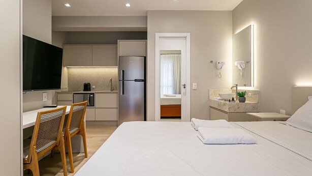 Una habitación de hotel moderna con cama, escritorio con dos sillas, televisor, zona de cocina con refrigerador y baño con espejo y lavamanos.