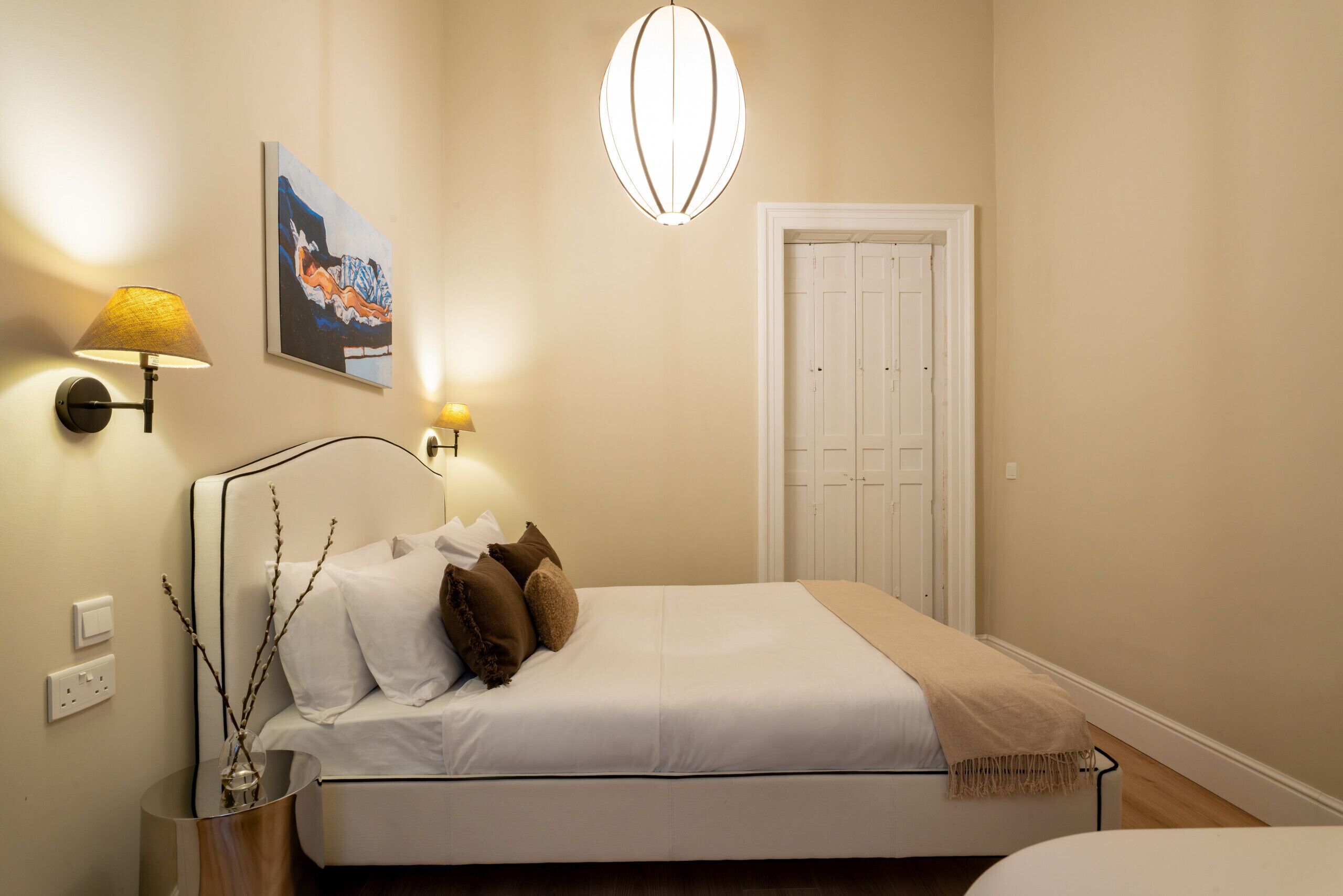 Een slaapkamer met een bed, nachtlampjes, een schilderij, een deur en een wandlamp.