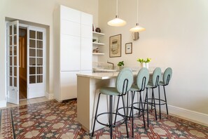 A kitchen with a tiled floor, white cabinets, a bar area with stools, pendant lights, and a door leading to another room. - Stylish 2 bedroom flat with amazing views LDEM1-5 (Valletta)