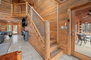 Interno di una baita in legno con una scala, un camino, una zona pranzo e un'ampia finestra.