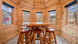 A wooden cabin interior with a round table and four stools, large windows, and a tiled floor. - Large Hot Tub | Pool Table | Playset | Sleeps 22 (Broken Bow)