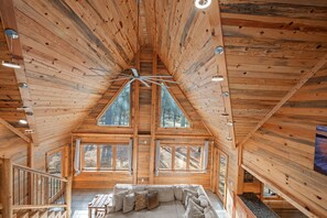 A wooden cabin interior with a sloped ceiling, large windows, and a central ceiling fan. - Large Hot Tub | Pool Table | Playset | Sleeps 22 (Broken Bow)