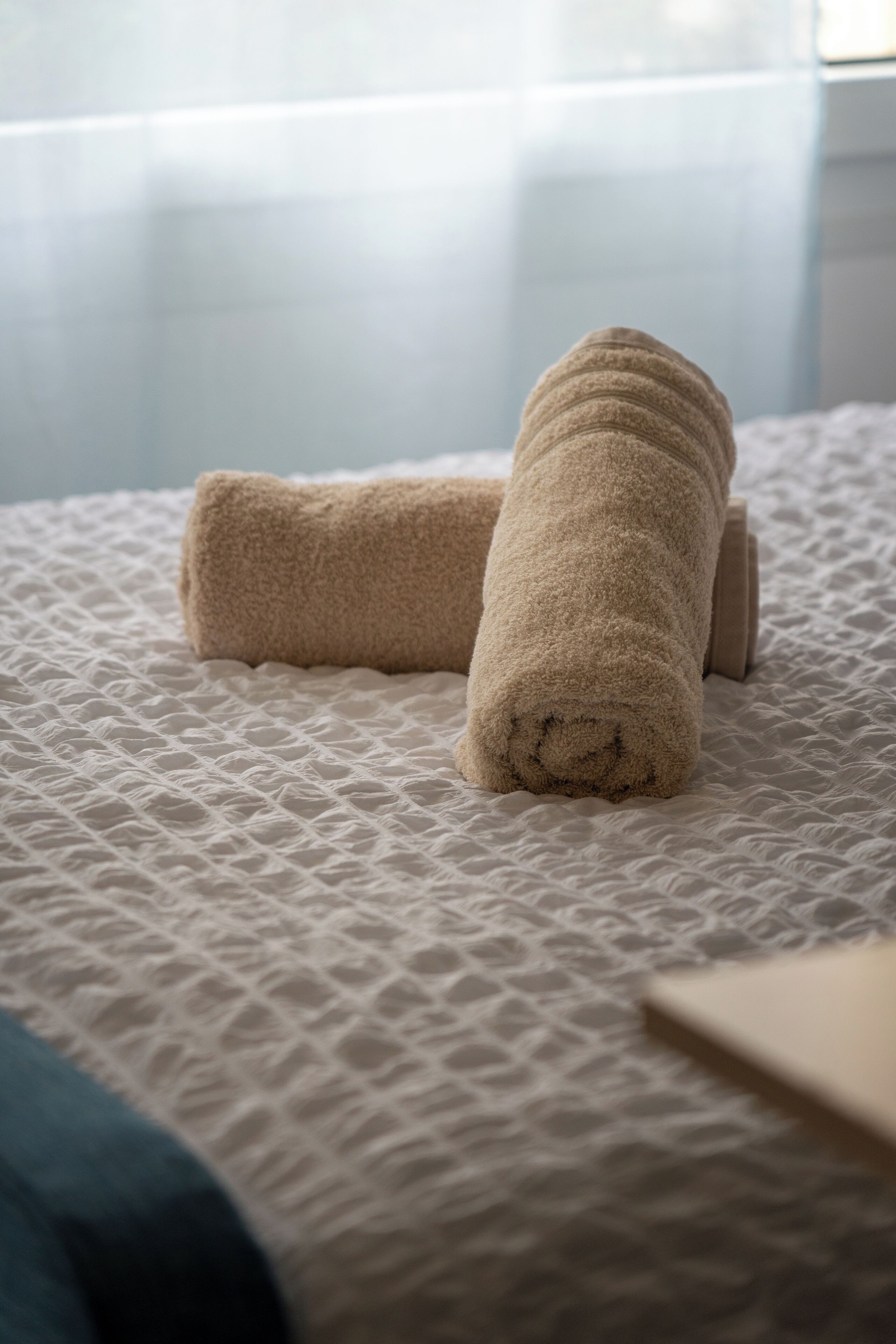 A bed with a textured white cover and two rolled towels.