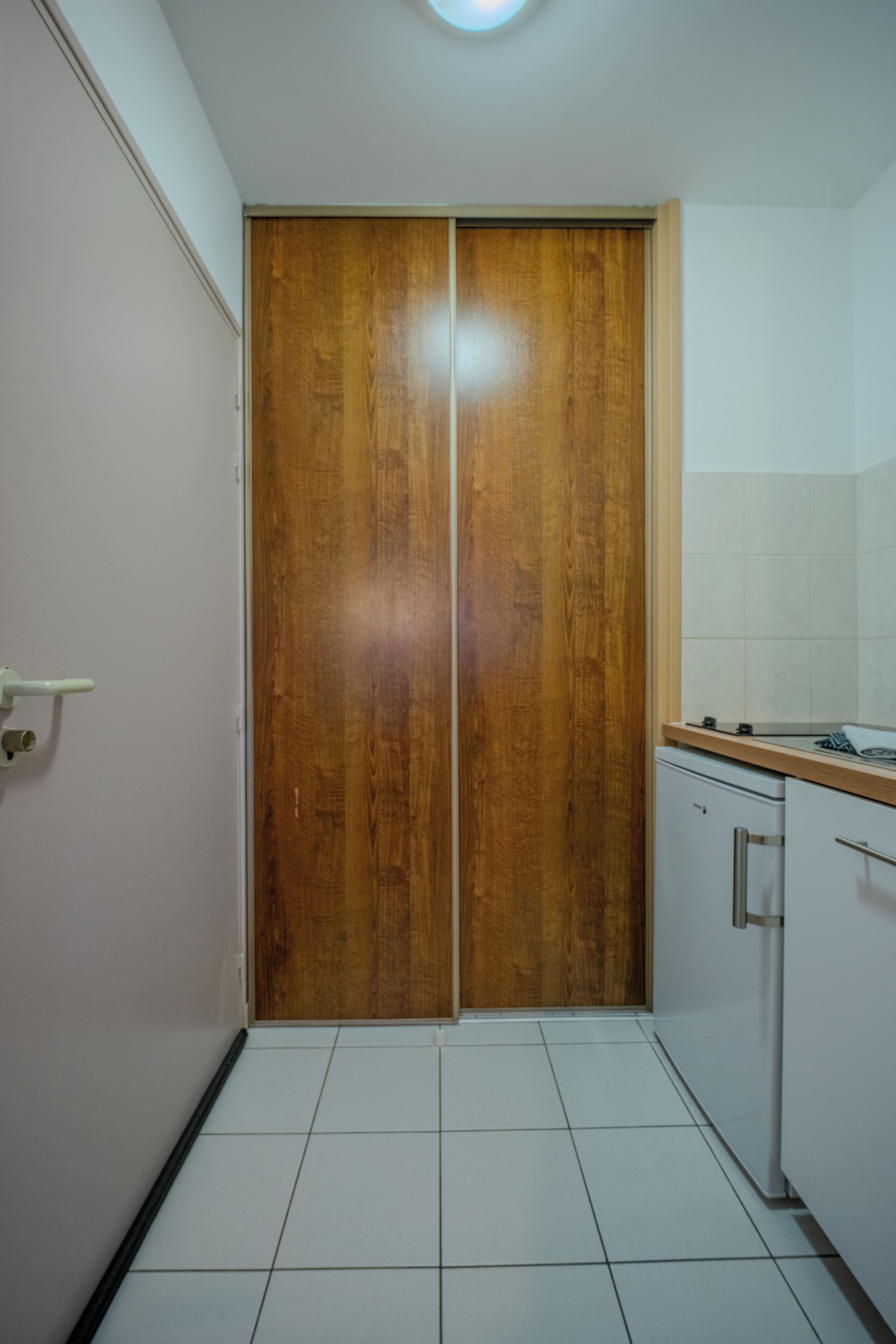 A compact kitchen with a wooden door, white tiled floor, and a small refrigerator.