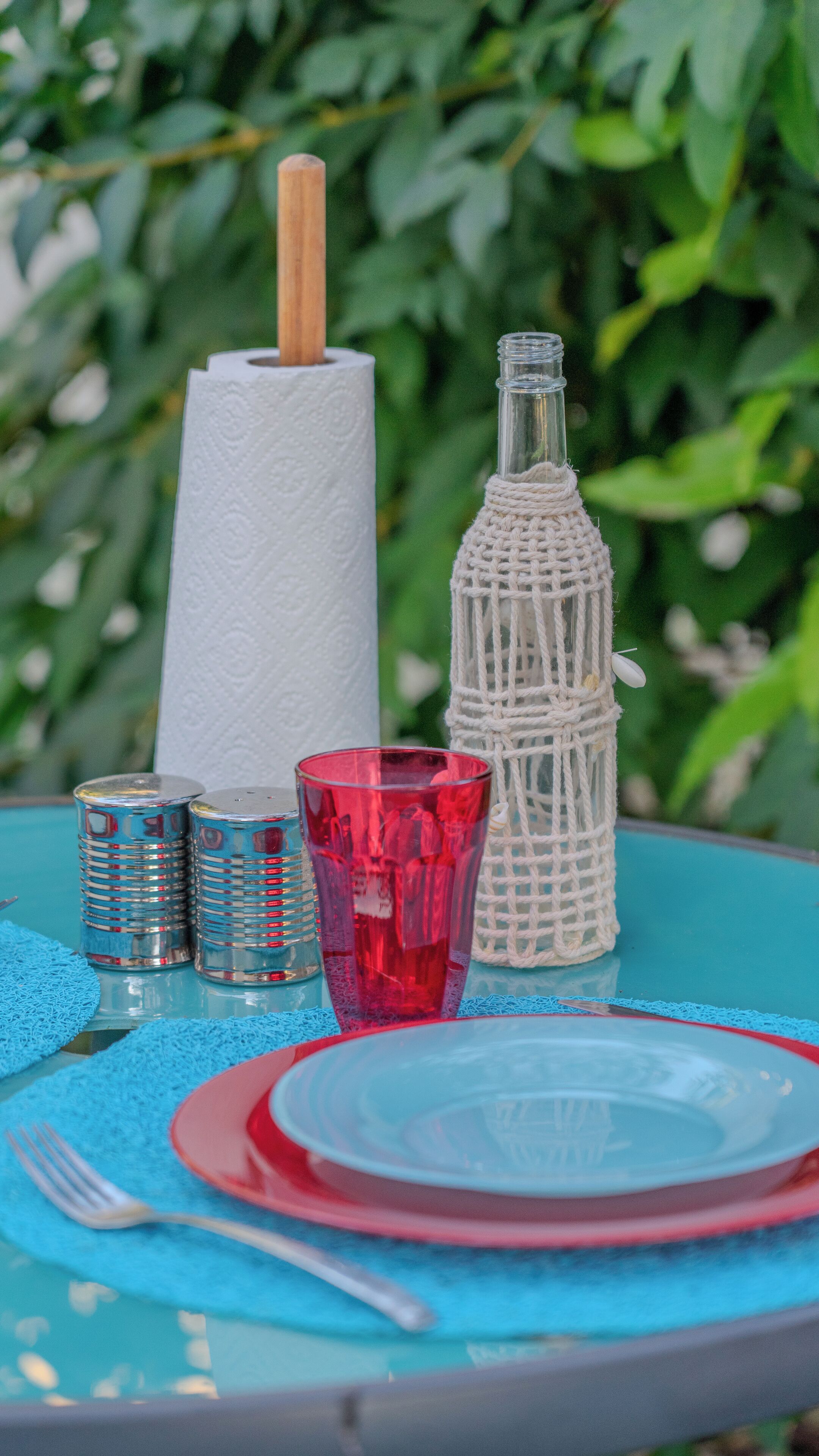 A table set with a red plate, a red glass, a silver candle holder, a white napkin holder, and a woven bottle.