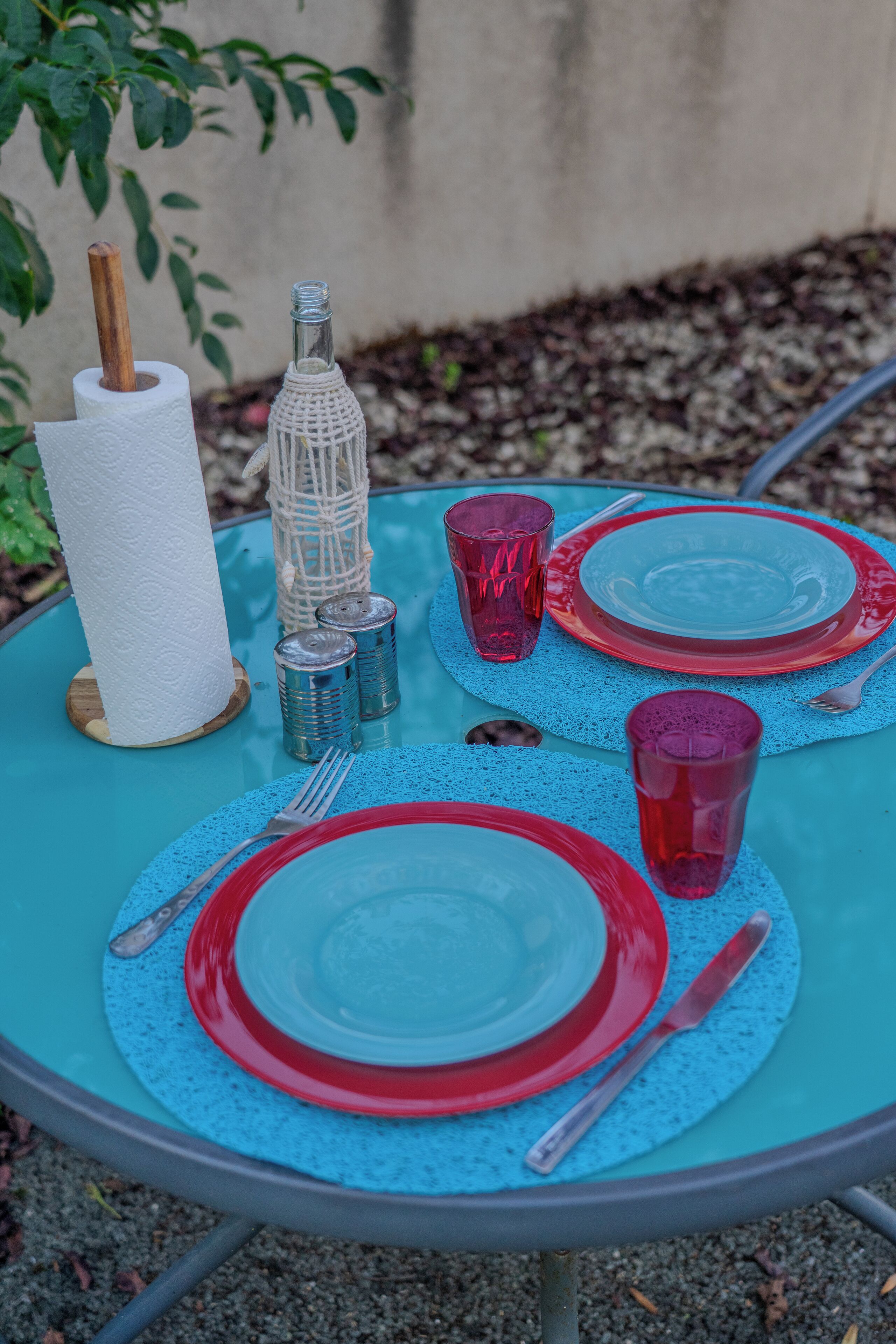 Une table dressée à l’extérieur, avec de la vaisselle en verre rouge et transparent, un porte-serviettes en papier et une bouteille décorative.