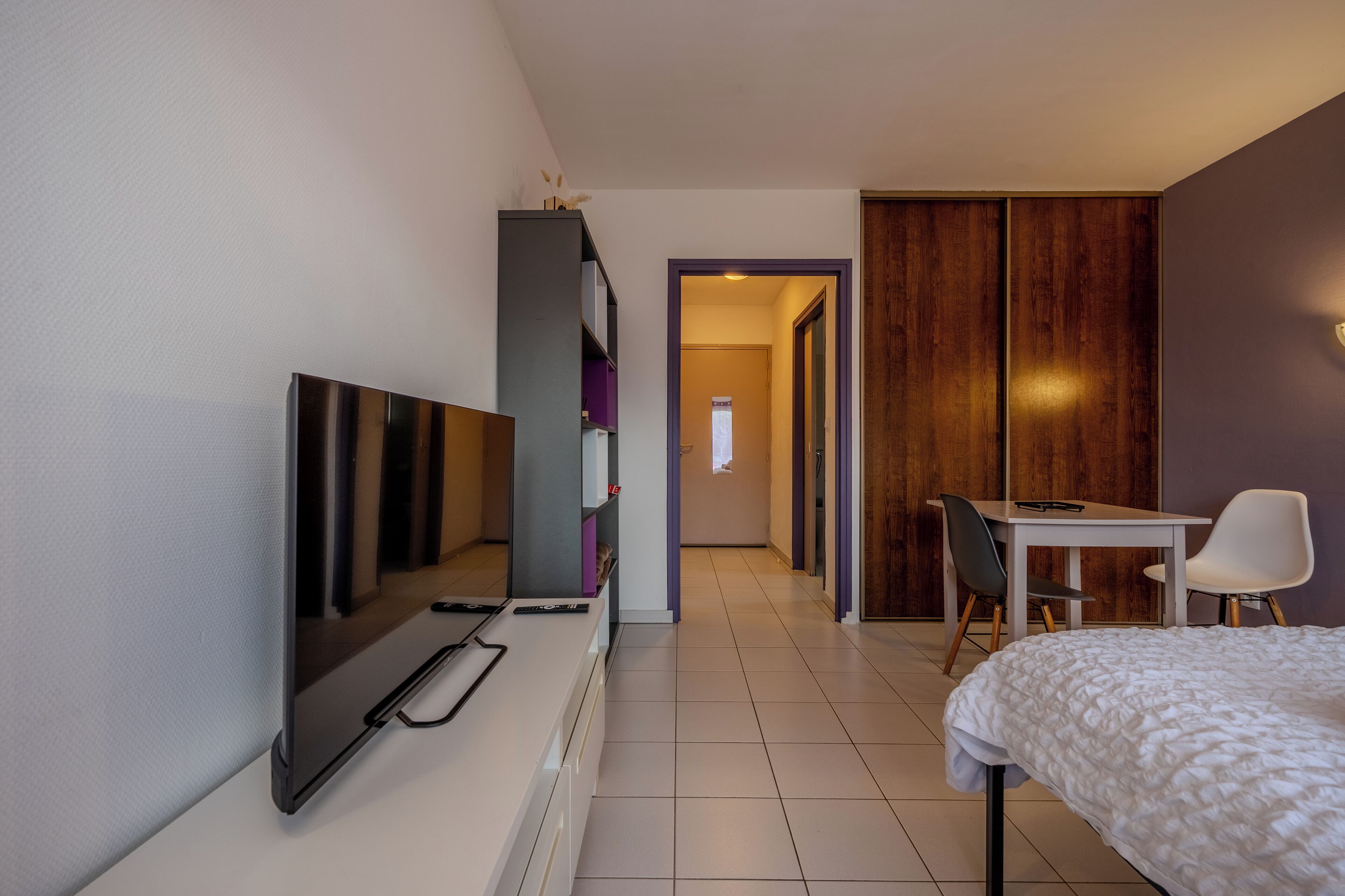 A modern hotel room with a bed, a desk, a chair, a TV, and a wardrobe.