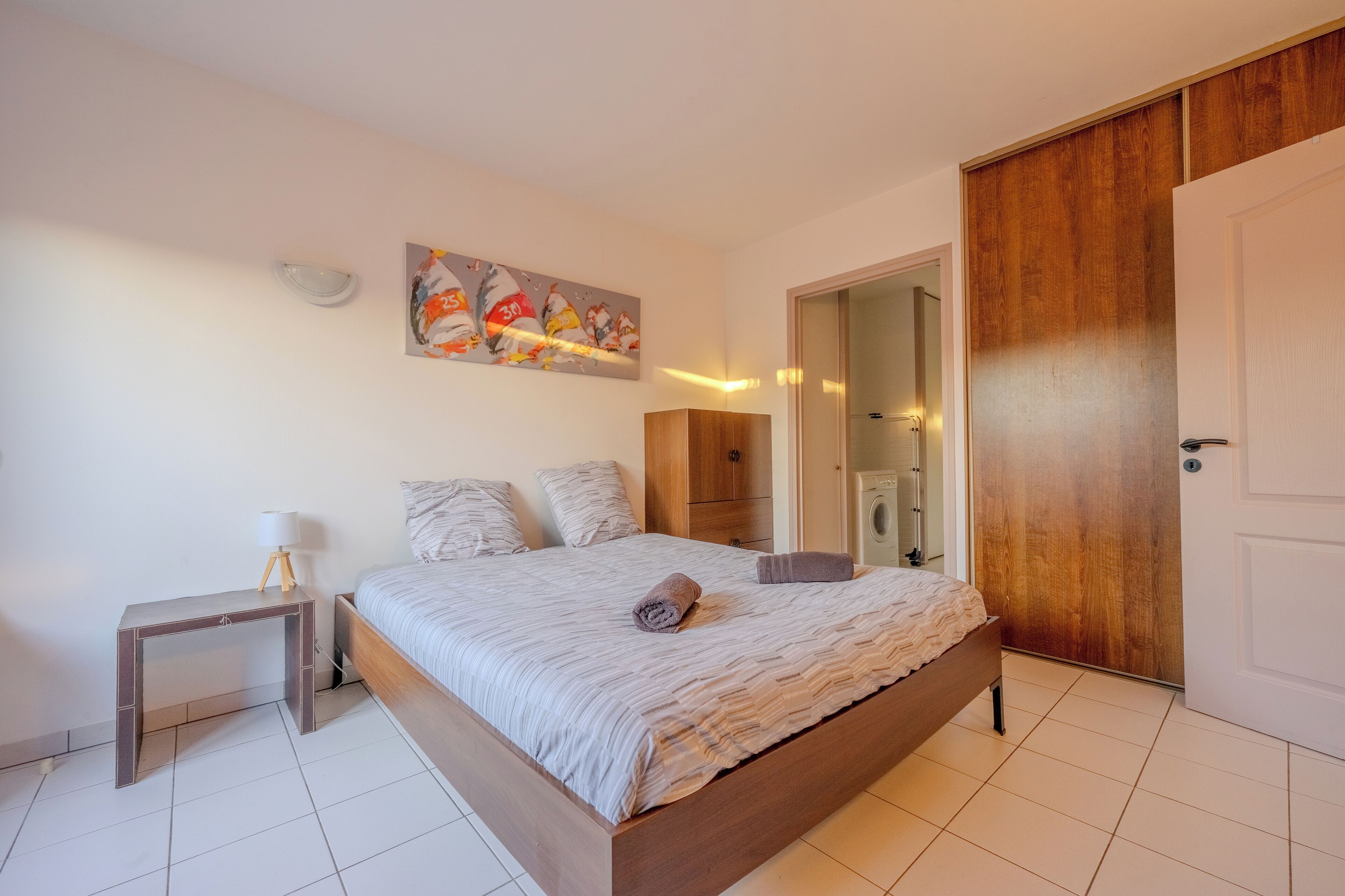 Un dormitorio con cama, mesita de noche, lámpara y un cuadro en la pared.