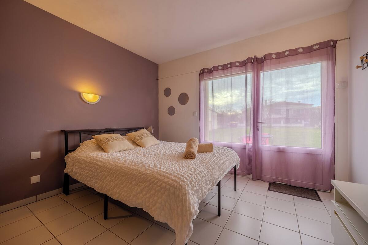 A bedroom with a bed, a wall-mounted light, a window with curtains, and a tiled floor.